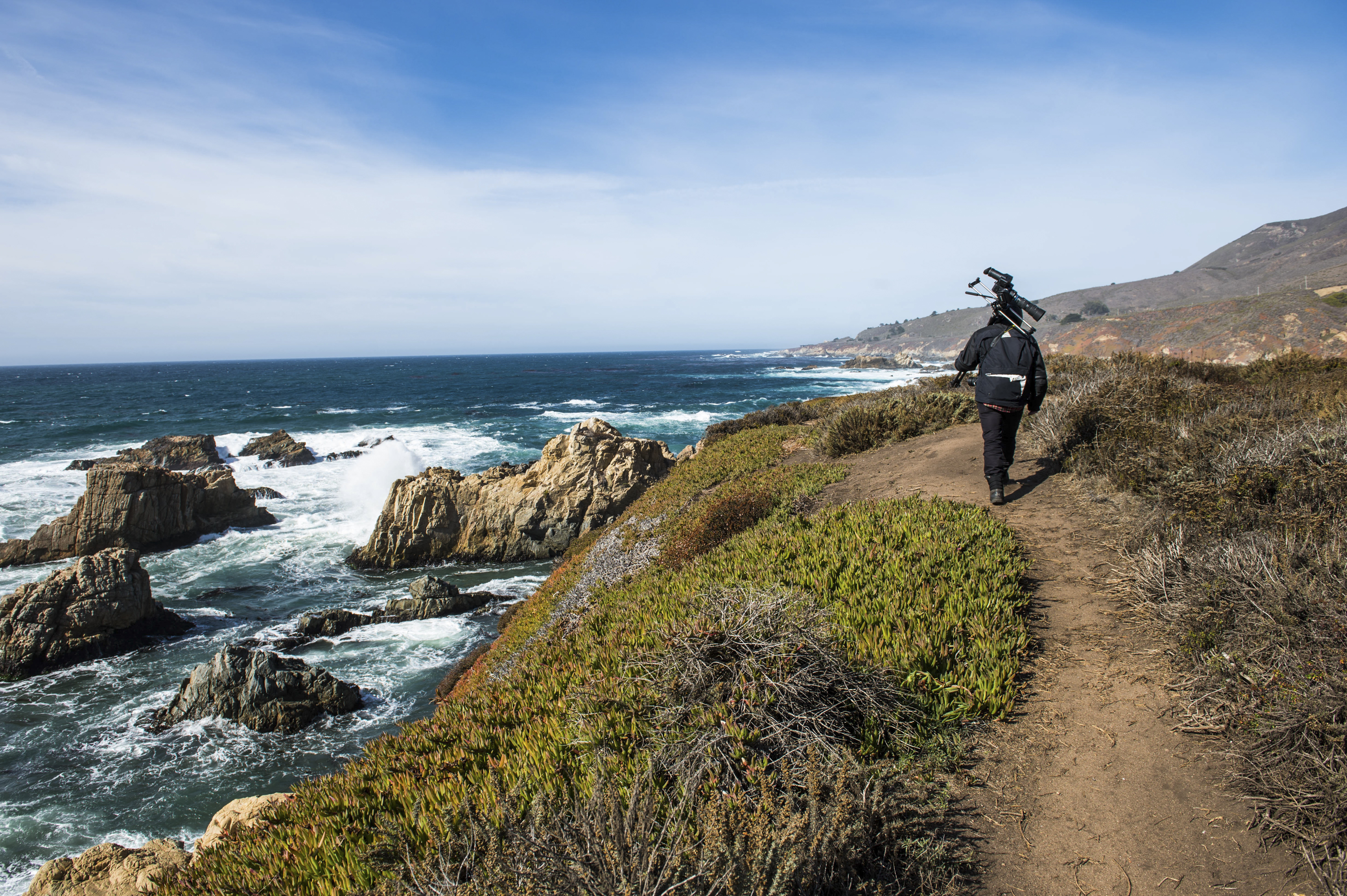 Camera operator hiking a rugged Big Sur coastal trail above the surf
