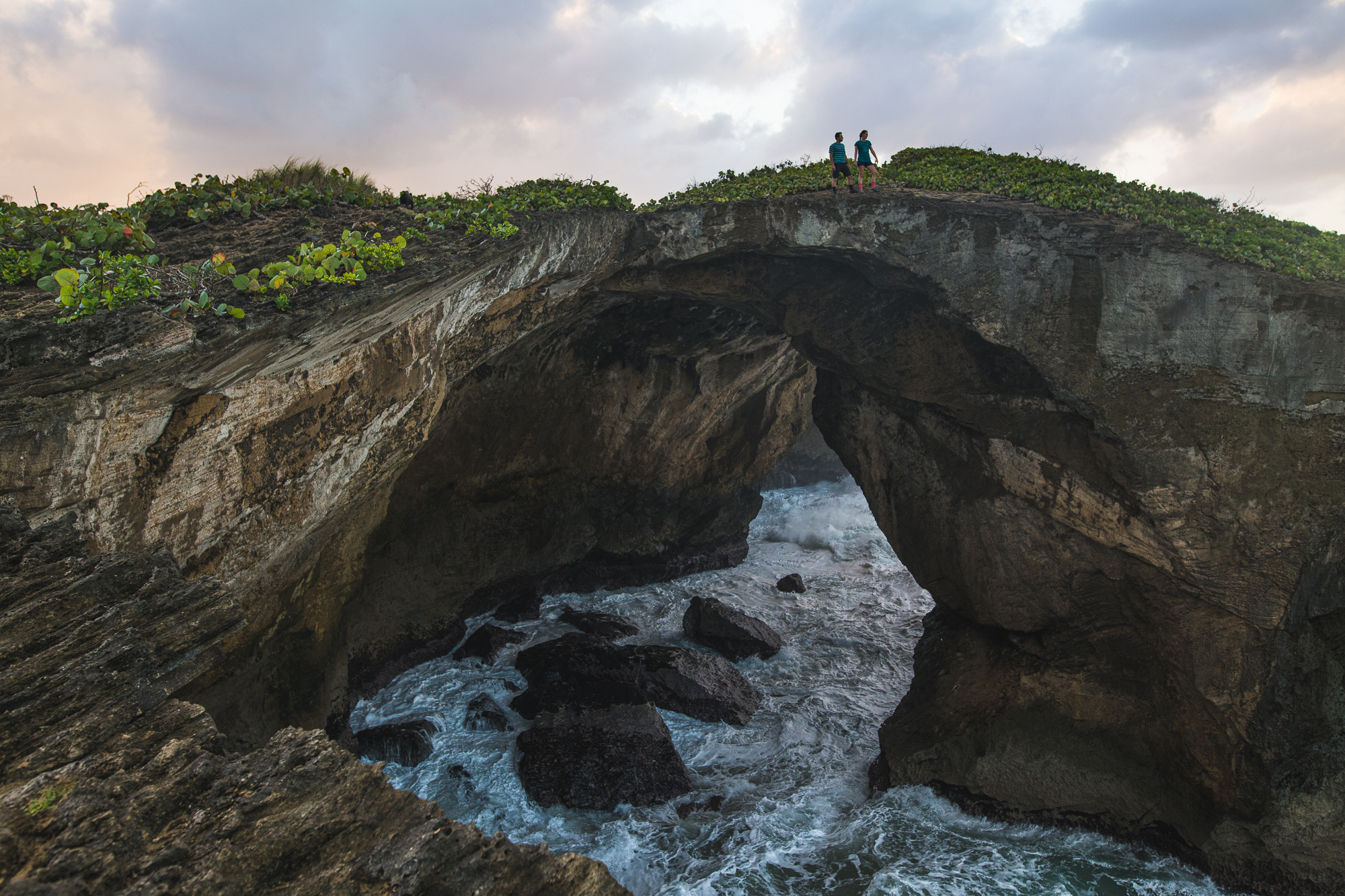 Two people standing atop a dramatic sea arch at dusk